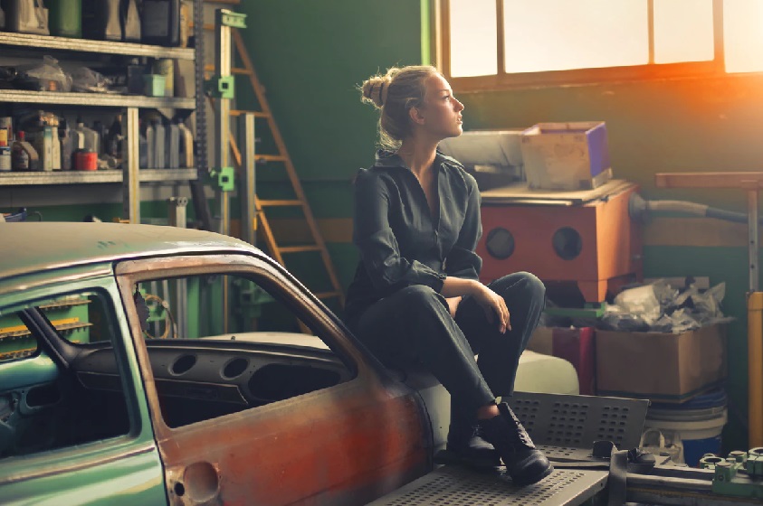 ガレージの古ぼけた車に腰かけて、窓から入る夕日を見ながら考え事をする女性